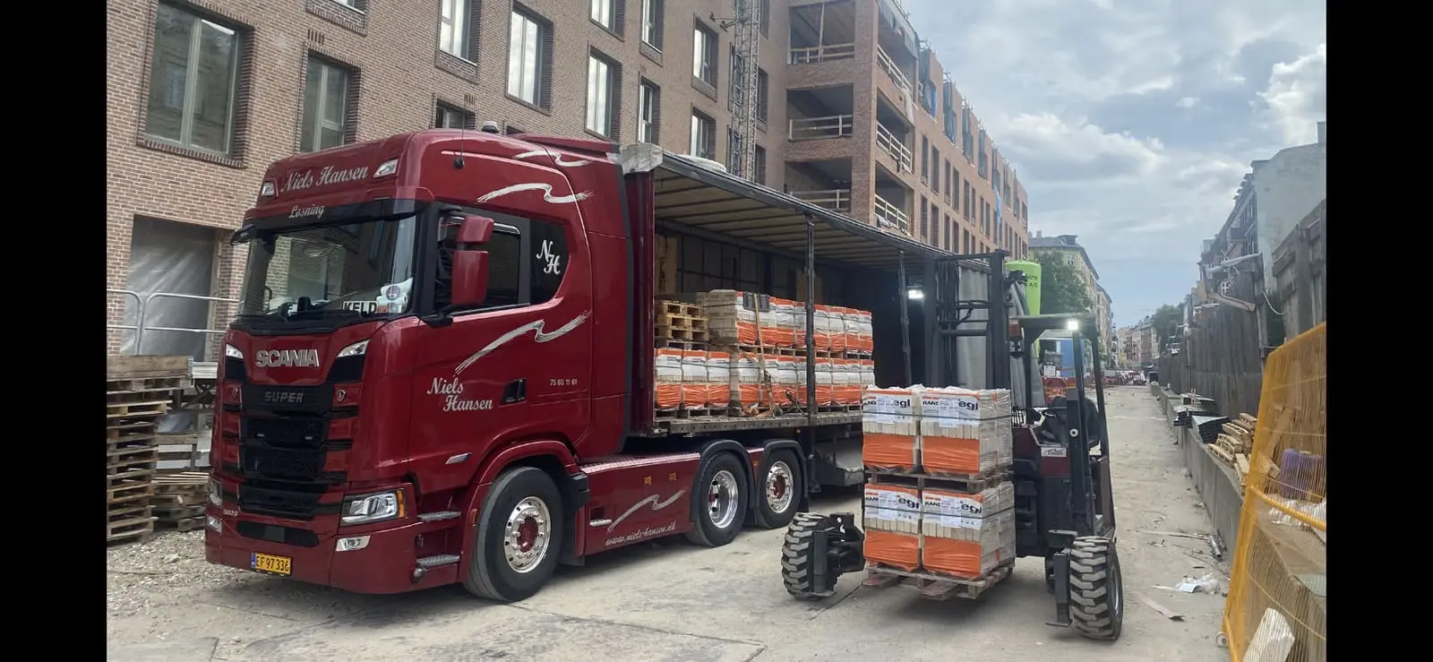 Medbringertruck kører to paller byggematerialer af trailerens åbne lad på en byggeplads i en smal gade