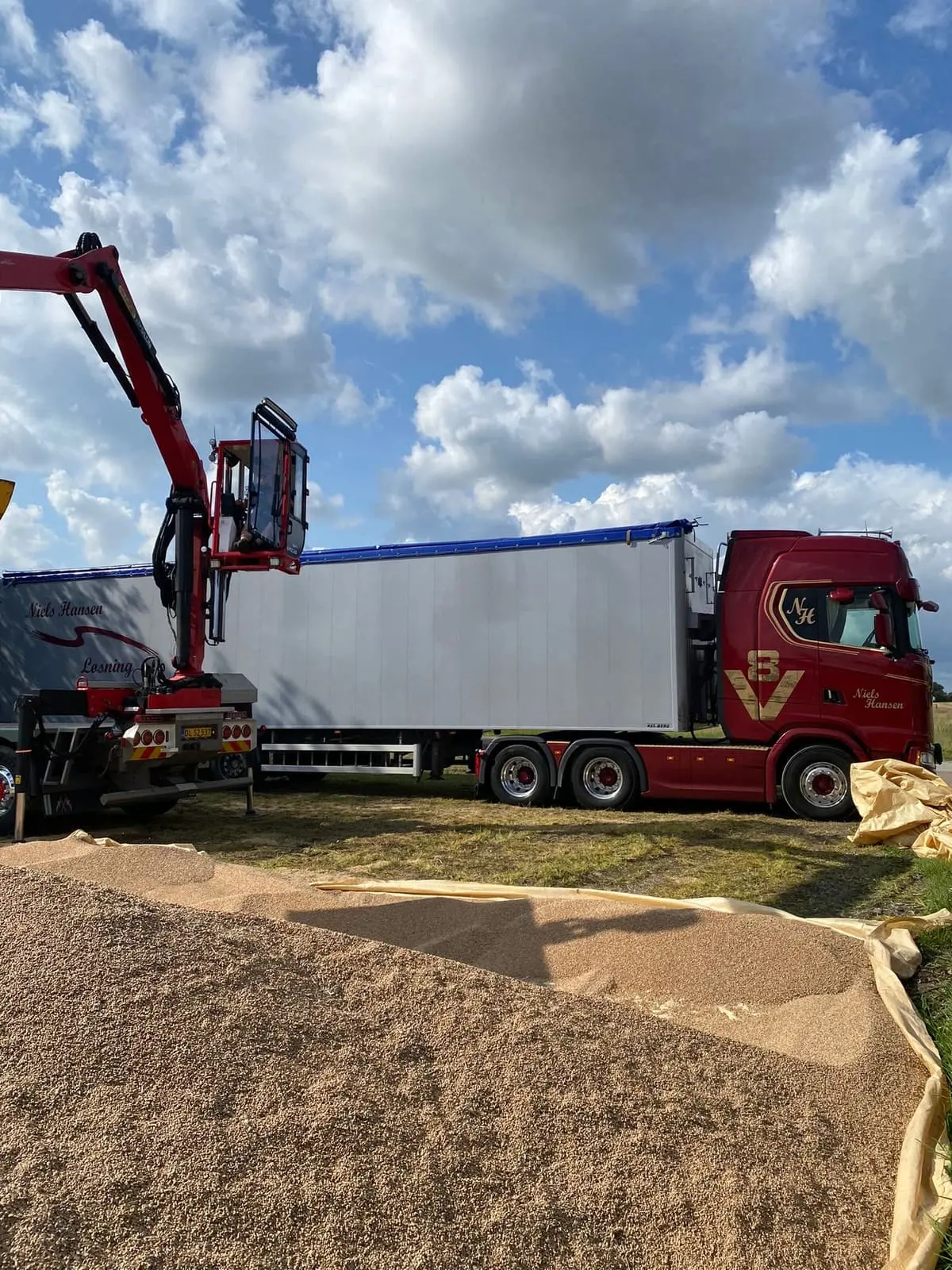Lastbil med hvid bulktrailer holder ved en stor dynge gulligt materiale på marken