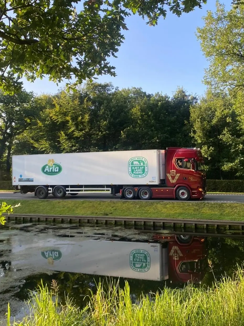 Lastbil med hvid kassetrailer spejlet i vandet langs vejen