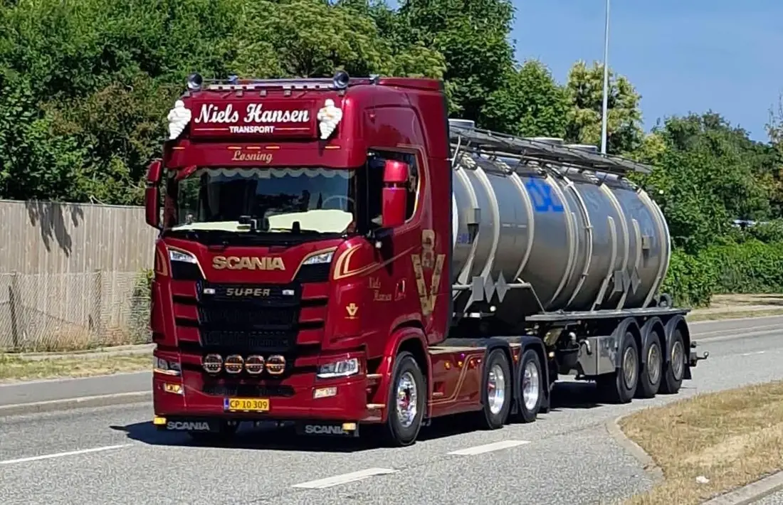 Rød lastbil med blank ståltank på vejen mellem grønne træer