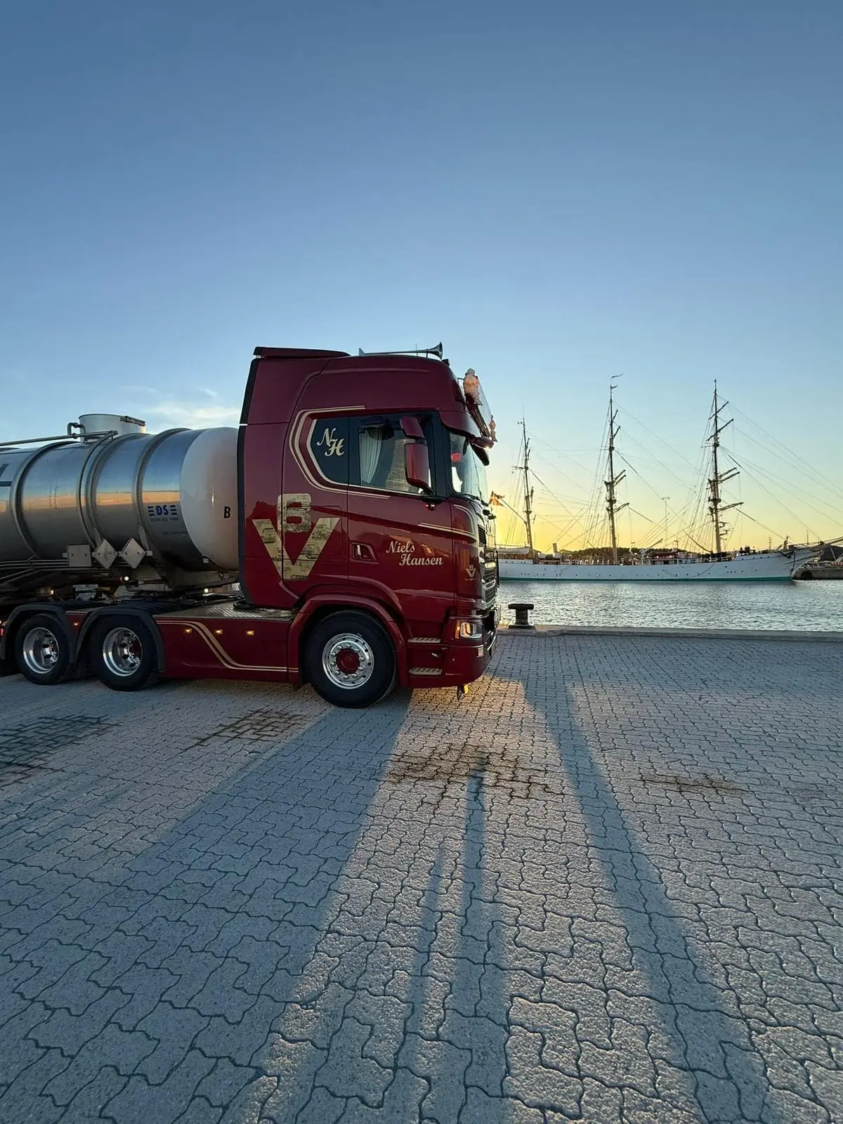Lastbil med tanktrailer på kajen i aftensol med et sejlskib i baggrunden