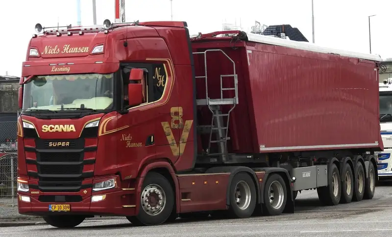 Rød lastbil med firakslet tippertrailer på vej langs et industriområde