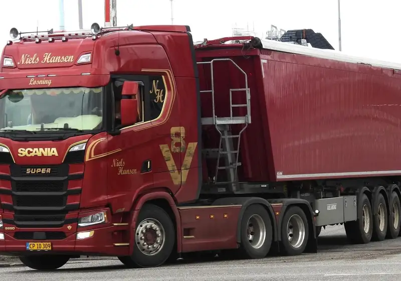 Rød lastbil med firakslet tippertrailer på vej langs et industriområde