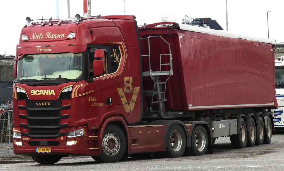 Rød lastbil med firakslet tippertrailer på vej langs et industriområde
