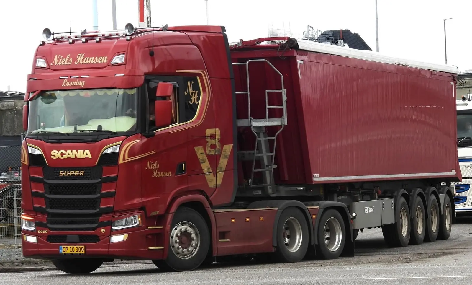 Rød lastbil med firakslet tippertrailer på vej langs et industriområde