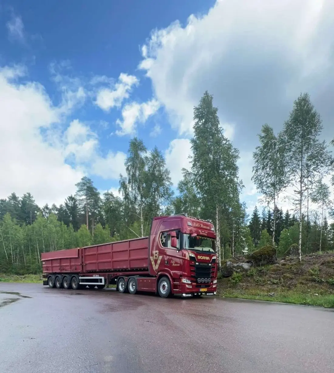 Rød lastbil med højsidet tiptrailer parkeret på våd asfalt ved skoven