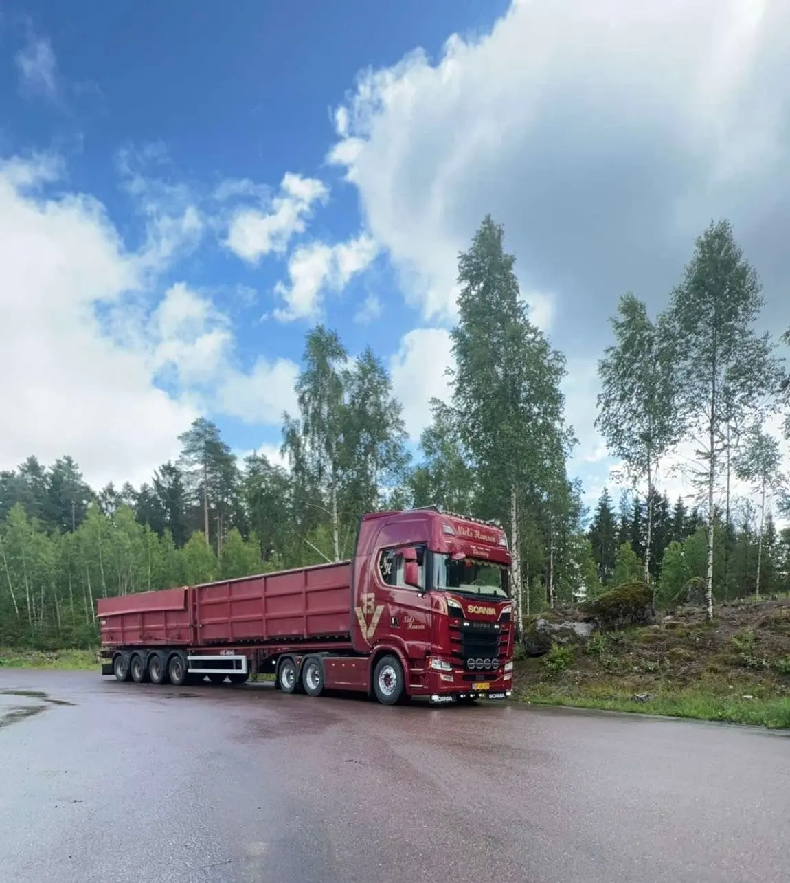 Rød lastbil med højsidet tiptrailer parkeret på våd asfalt ved skoven