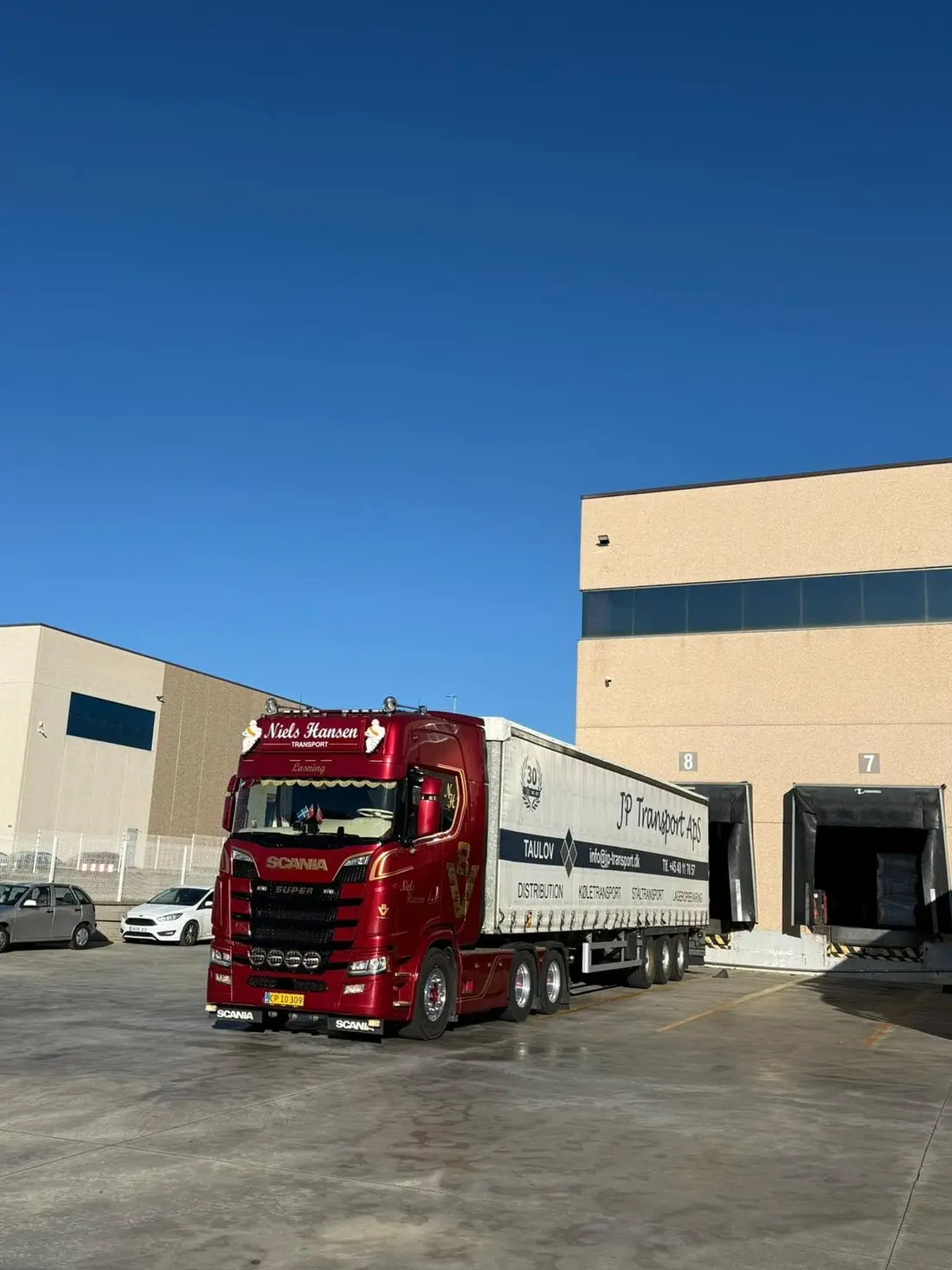 Rød lastbil med gardintrailer holder ved lagerportene i skarp sol