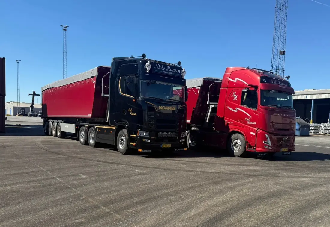 En sort og en rød lastbil holder side om side med tippertrailere i solskin